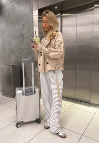 Trench beige chiaro, pantaloni a righe a gamba larga, valigia rigida argentata con manico estensibile e sneakers con motivo. Tiene una bevanda.