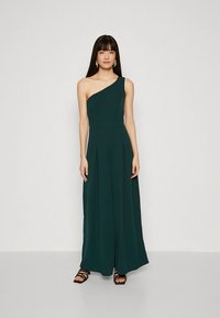 Egyszárú sötétzöld overall széles szárakkal, redőzött előlappal és sima anyagból. A modell fekete pántos szandált és minimális ékszereket visel.