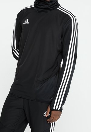 Person iført et sort Adidas sportsoutfit med hvide tre-stribede detaljer på ærmer og bukser, stående mod en ensartet baggrund.