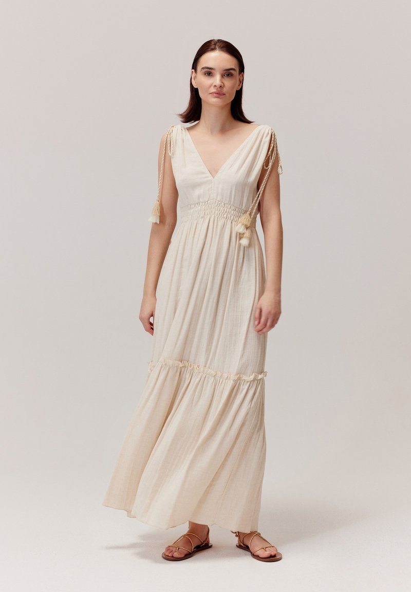 Długa, kremowa maxi sukienka z dekoltem w kształcie litery V, gorsetem marszczonym i wiązanymi ramiączkami ozdobionymi frędzlami. Lekki materiał; warstwowy falbany u dołu spódnicy.
