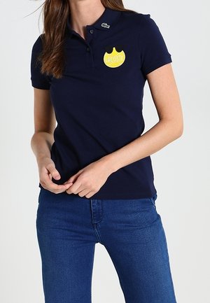 Femme portant un polo Lacoste bleu marine avec un logo de balle de tennis jaune et un jean bleu, debout les mains jointes à la taille.