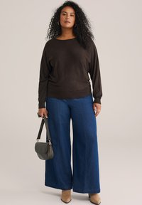 Hnědý lehký svetr s lodním výstřihem, zkombinovaný s modrými volnými denimovými kalhotami a malou zelenou kabelkou. Měkká textura a uvolněný střih.