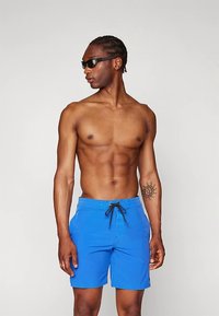 Modré plavky s elastickým pásom a bočnými vreckami. Hladká tkanina, dĺžka nad kolená, v kombinácii s čiernymi slnečnými okuliarmi.