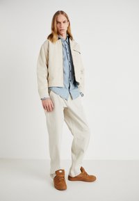 Giacca beige corta sopra una camicia a righe blu, abbinata a pantaloni crema e scarpe da ginnastica marroni. Tessuto liscio, vestibilità rilassata e accenti minimi.
