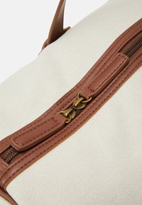 Torba canvas w kolorze kremowym z brązowymi skórzanymi akcentami i mosiężnym zamkiem, podkreślająca przeszycia i fakturę powierzchni.