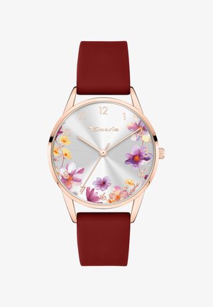 Rosaguld ur med en sølvskive med blomsterdesign, der har et rødt silikonerem. Skiven viser timer fra 1-12 markeret i guld.