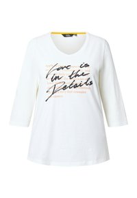 Camicia bianca a maniche lunghe con scollo a V e testo "L'amore è nei dettagli" in nero e con una scritta arancione tenue sul davanti.