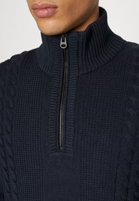 Marinblå stickad tröja med hög krage och halvdragkedja, med kabelstickningsdetaljer på sidorna för textur.