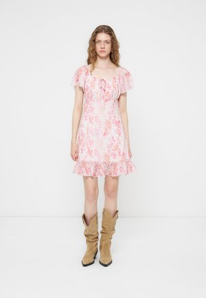 Jeune femme aux cheveux ondulés portant une robe courte rose à fleurs avec des manches volantées et des bottes cowboy marron clair décontractées, debout sur un fond blanc.