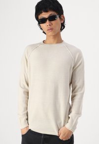 Pull en beige texturé avec des rayures, col rond et manches longues ; porté sur un pantalon noir, le modèle pose les mains dans les poches.
