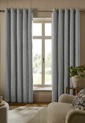 Hellblaue gemusterte Vorhänge hängen offen vor einem Fenster mit Blick auf einen Garten, daneben steht ein beiges Sofa und eine Topfpflanze auf einem Beistelltisch.