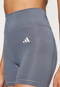 Pantaloni scurți gri, cu talie înaltă, confecționați dintr-un material elastic, cu logo-ul alb Adidas pe partea stângă și cusături netede, plate.
