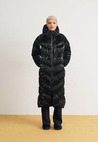 Svart, lång pufferjacka med ett chevronmönster, hög krage och dragkedja fram. Tillverkad av glänsande, kviltat material. Modell bär svarta byxor och skor.