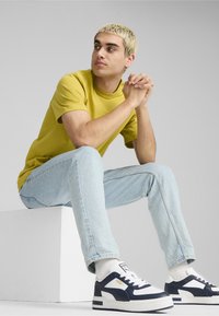 Dzeltena īsām piedurknēm t-krekls, gaiši zili džinsi un tumši balti sporta apavi, sēž uz balta bloka, rokas saliktas kopā, neitrāla fona.