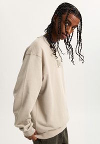Beige sweatshirt with a crew neck and relaxed fit, featuring a small embroidered logo patch on the chest and ribbed cuffs.