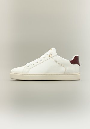 Baskets basses blanches avec lacets blancs, semelle crème, panneau latéral texturé et patch rouge foncé au talon sur fond gris neutre.