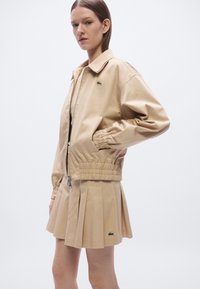 Beige bomberjack met ritssluiting en elastische taille, gecombineerd met een bijpassende geplooide rok. Lacoste-logo op jas en rok. Soepele textuur.
