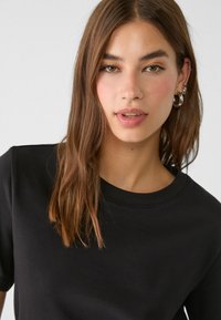Sort T-shirt med rund halsudskæring og korte ærmer, lavet af blødt stof. Modellen har langt, glat hår og minimal smykker.