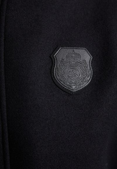 Must pikk villane mantel, millel on tekstuurse nahast emblem koos krooniga ja keeruka detailidega, õmmeldud kangale täiendava rõhutamise jaoks.