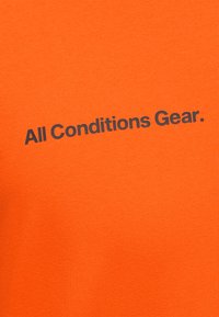 Φωτεινό πορτοκαλί ύφασμα με μαύρο κείμενο "All Conditions Gear." τυπωμένο διαγώνια στο κέντρο.