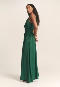 Dlouhé zelené šaty z lehké látky, s přiléhavým živůtkem, staženým pasem na šňůrku a splývavou sukní s minimem kovových doplňků.