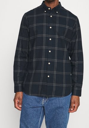Mand iført en langærmet sort skjorte med gråt ternet mønster og blå jeans, står mod en ensfarvet lys baggrund.