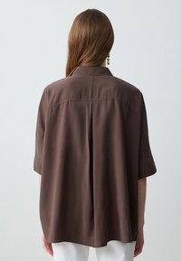 Braunes kurzärmeliges Hemd mit lässiger Passform, ausgestattet mit einem Rückenschlitz und weichem Material, von hinten mit weißen Hosen gezeigt.