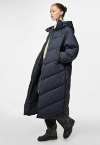 Námořnický modrý prošívaný kabát s kapucí, s dlouhým, oversized střihem, prošívanými částmi a dvěma bočními kapsami. Nosený přes lehký pletený svetr a džíny.