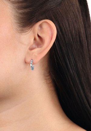 Pequeño pendiente de aro plateado llevado en la oreja izquierda de una persona con cabello liso castaño oscuro visible detrás de la oreja.