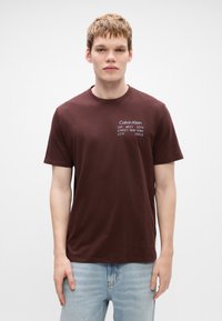 Camiseta de algodón marrón con cuello redondo, mangas cortas y detalles en texto blanco que dicen "Calvin Klein" y la dirección. Lleva puesta con jeans de color azul claro.