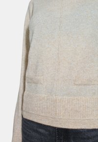 Maglione beige chiaro con lavorazione a maglia testurizzata, orlo a coste e due tasche frontali. Presenta un design a taglio dritto e dettagli delle cuciture in vista.