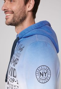 Felpa con cappuccio blu a gradiente con coulisse, dotata di una stampa grafica frontale e un emblema sulla manica sinistra. Morbido al tatto e dal design casual, adatta per essere indossata a strati.