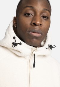 Witte fleece jas met een hoge kraag, voorzien van zwarte trekkoordaccenten en een ritssluiting, met een getextureerd, zacht oppervlak.