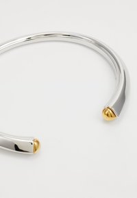 Pulsera de metal de tono plateado con un diseño suave y curvado. Presenta dos extremos redondeados de color dorado, que destacan un acabado metálico en contraste.