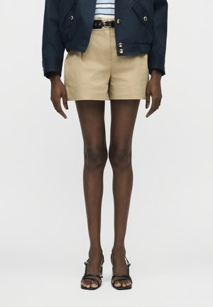 Μοντέλο που φοράει μπεζ σορτς ψηλής μέσης με μαύρη ζώνη, μπλε ναυτικό σακάκι με χρυσά κουμπιά, ριγωτό τοπ, μαύρα διάφανα καλσόν και μαύρα σανδάλια με τακούνι.