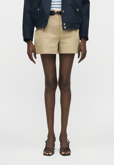 Mannequin portant un short beige taille haute avec ceinture noire, veste bleu marine à boutons dorés, haut rayé, collants noirs transparents et sandales à talons noires.
