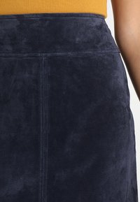 Suedeskjol i marinblått med en slät textur och rena sömmar. Har en rak skärning och en hög midja, delvis synlig högst upp.