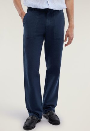 Uomo che indossa pantaloni blu navy a gamba dritta, mocassini neri e camicia azzurra a righe infilata, in piedi con le mani rilassate.