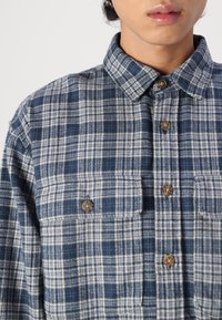 Camisa de franela de cuadros en azul marino y azul claro con cuello abotonado, dos bolsillos en el pecho y botones marrones. Tejido texturizado con un patrón de cuadros.