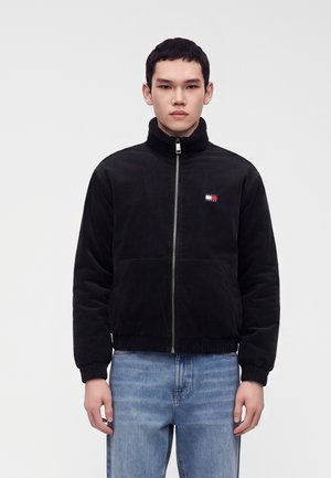 Giovane uomo che indossa una giacca in pile nera con zip e un piccolo logo di bandiera, e jeans azzurri, in piedi davanti a uno sfondo bianco semplice.