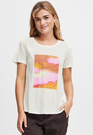 Donna che indossa una maglietta color crema con stampa astratta rosa e arancione, pantaloni neri e orecchini a cerchio dorati, sorridente con una mano in tasca.