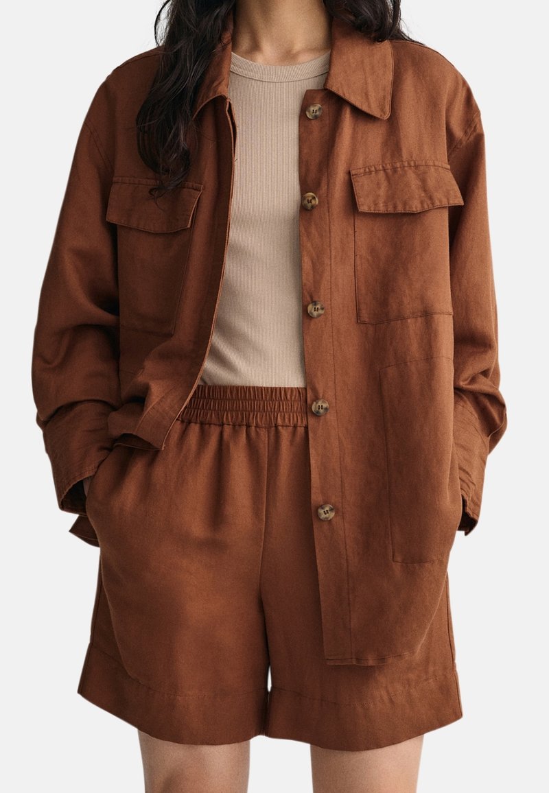 Femme portant une veste-chemise couleur rouille à boutons avec deux poches poitrine et un short assorti taille élastique sur un haut côtelé beige.