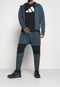 Tuta sportiva blu con rifiniture nere; giacca con cappuccio full zip sopra una maglietta nera con logo bianco, abbinata a pantaloni jogger coordinati e scarpe nere.