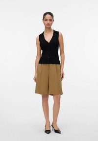 Zwart geribbeld mouwloos topje met een V-hals en knopen, gecombineerd met tan geplooide shorts en zwarte puntige hakken. Eenvoudig design en kleuren.
