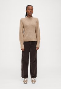 Maglione a costine beige con collo alto, abbinato a pantaloni in corduroy a gamba larga marrone scuro e sneakers beige chiaro.