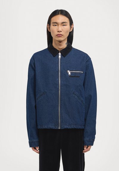 Indigo denim jacket with a black corduroy collar, front zip closure, a chest pocket with a zipper, and angled side pockets.
