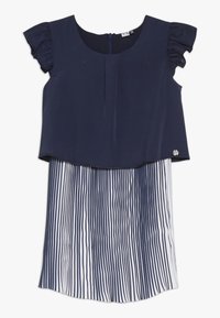 Námořnická modrá blůza s volnými rukávy pokrývá bílou sukni s vertikálními námořnickými pruhy. Hladká textura s kulatým výstřihem.