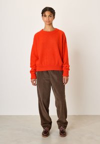 Une femme se tient devant un fond uni, portant un pull orange vif, un pantalon ample en velours côtelé marron et des chaussures à lacets marron foncé.