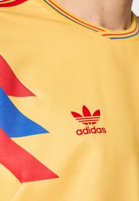 Maglietta gialla Adidas con motivi geometrici rossi e blu, con logo rosso sull'area sinistra del petto. Realizzata in tessuto liscio.