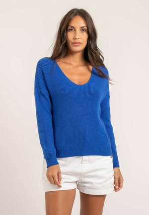 Femme aux longs cheveux noirs portant un pull bleu à col en V et un short blanc, debout devant un fond clair uni.